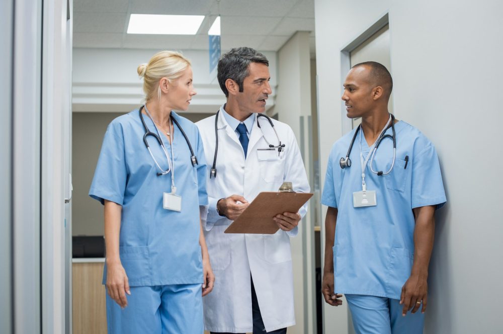 Mature doctor discussing with african male nurse in a hallway hospital. Doctor discussing patient case status with his medical staff after operation. Doctor holding clipboard while in conversation with nurse.